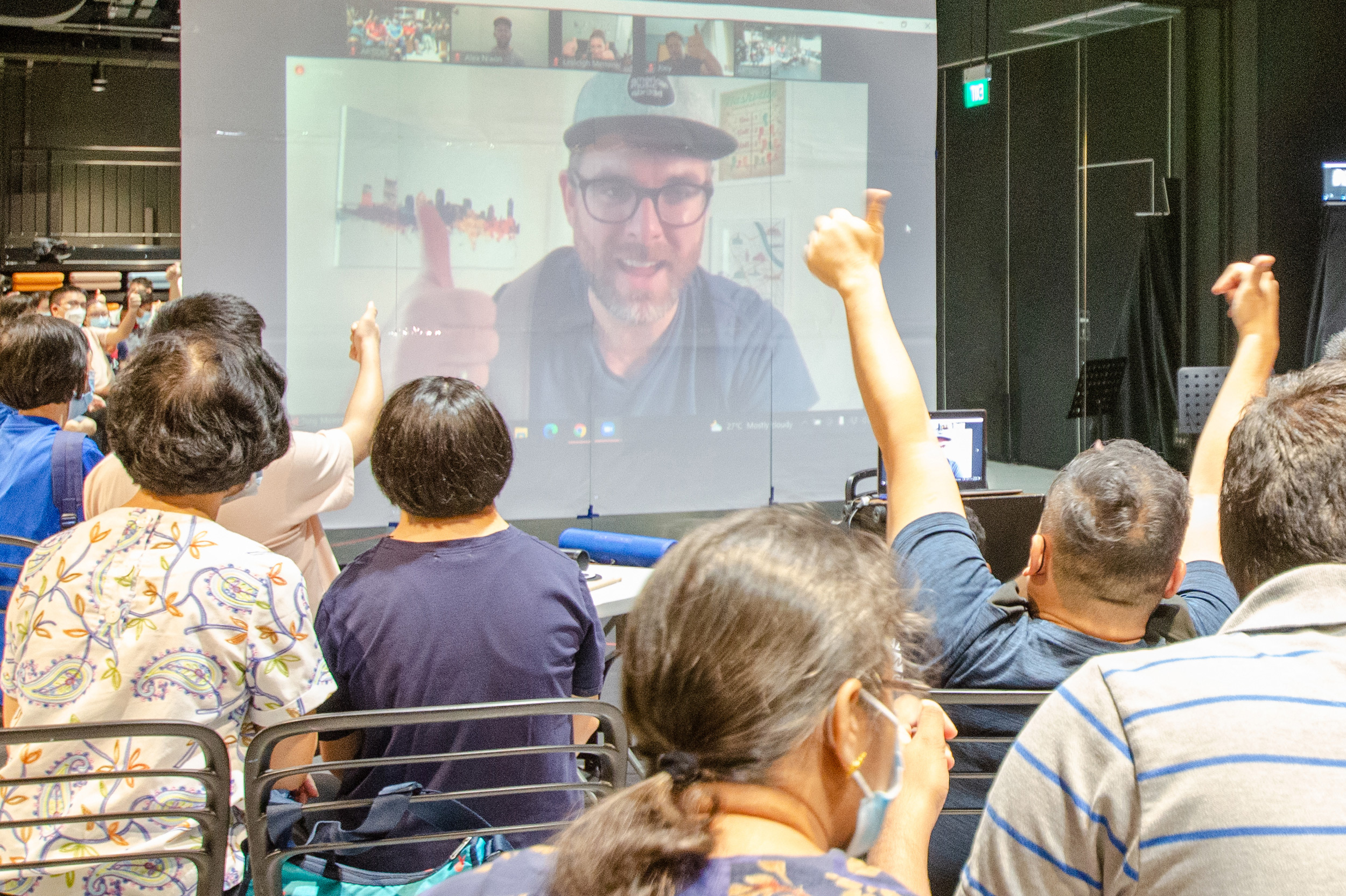 This image features Tony Memmel projected on a screen. He is making the thumbs-up sign and the participants are responding with raising their own hands in the hair with the thumbs-up sign.